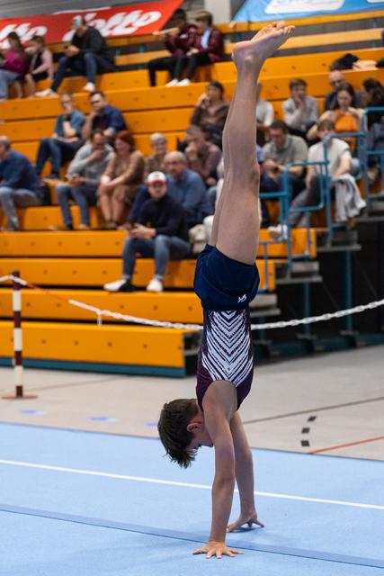 Young gymnast performs a straight handstand on blue mat, with spectators watching from yellow bleachers in background