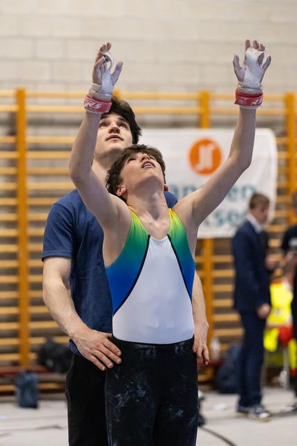 Gymnast in colorful leotard raises arms skyward while coach spots from behind in gymnasium