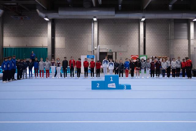 Multiple gymnastics teams standing on award podiums in an indoor sports hall during a medal ceremony celebration