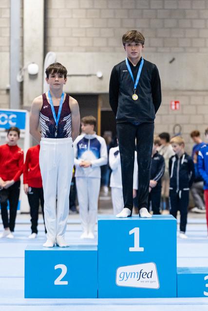 Two young gymnasts stand on award podium wearing medals, first place winner elevated above second place finisher