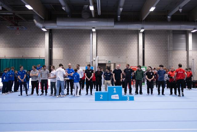 Large group of athletes and coaches gathered around a three-tier podium in an indoor sports facility with blue flooring