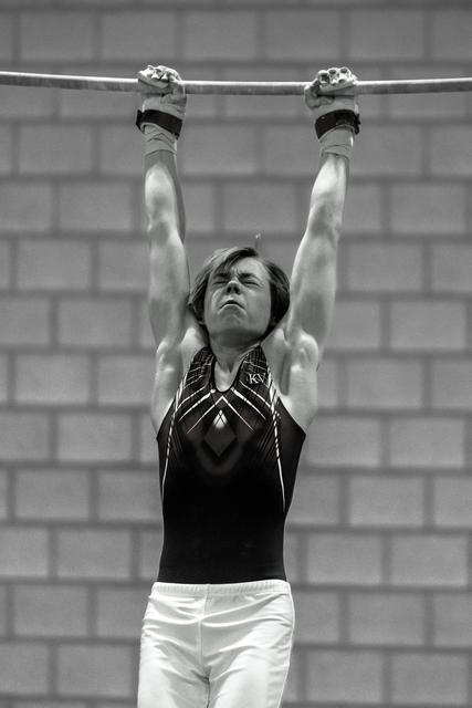 Male gymnast in black and white leotard focuses intently while gripping the horizontal bar in training facility