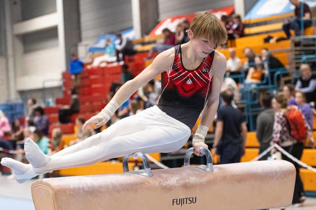 Young male gymnast performs scissors move on pommel horse, demonstrating strength and flexibility before crowd in gymnasium