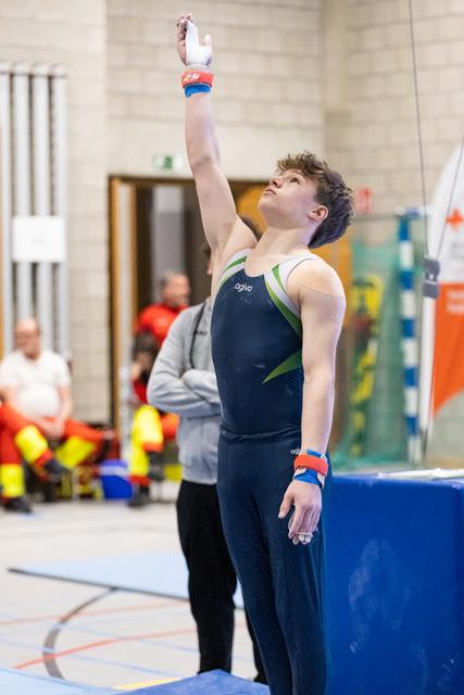 Male gymnast in navy leotard with arm raised in finishing salute pose, looking upward with focused expression in gym