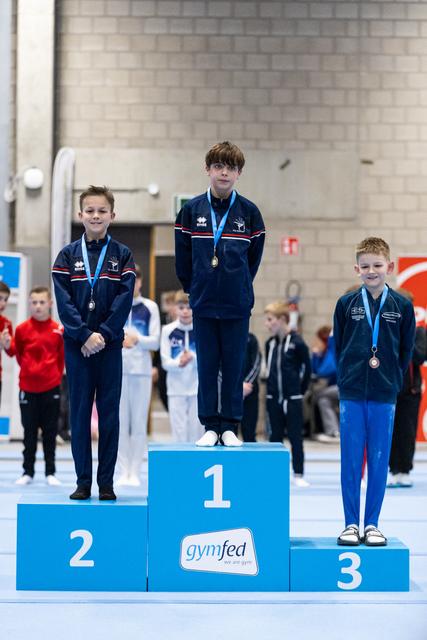 Three young gymnasts stand on award podium wearing medals, first place elevated on blue block marked '1' in sports hall