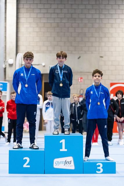 Three young male gymnasts stand on award podium wearing medals, first place in dark tracksuit, others in blue