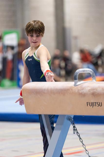 Young gymnast in navy and green leotard approaches pommel horse with focused expression during training session