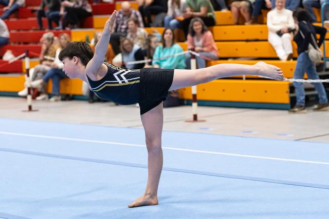 Young gymnast holds an arabesque balance position on floor exercise mat while spectators watch from bleachers