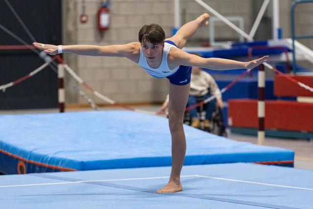 Young gymnast performs an arabesque balance on floor, arms extended gracefully, wearing light blue and navy leotard