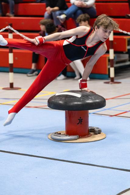 Young gymnast performs a horizontal balance hold on pommel mushroom, demonstrating strength and control in red uniform