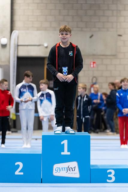 Young gymnast in black tracksuit stands proudly on first place podium holding medal, with competitors visible behind