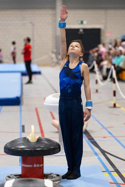 Young gymnast in blue leotard strikes finishing pose with raised arm beside mushroom training apparatus at gymnastics event