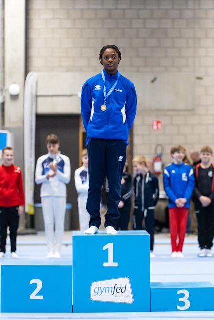 Young gymnast stands proudly on first place podium wearing blue tracksuit and gold medal, with competitors behind