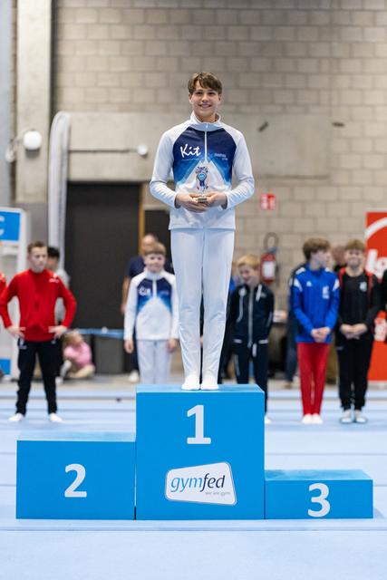 Young gymnast standing proudly on first place podium holding trophy, wearing USA warmup suit at gymfed event