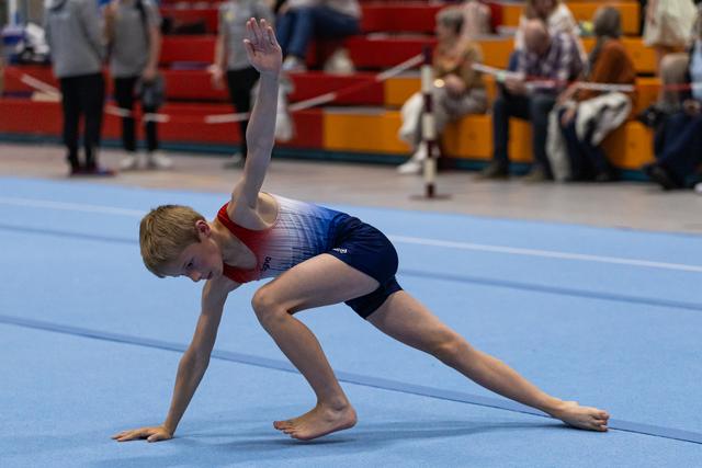 Young gymnast performs floor routine in artistic lunge pose with extended leg and raised arm during indoor event