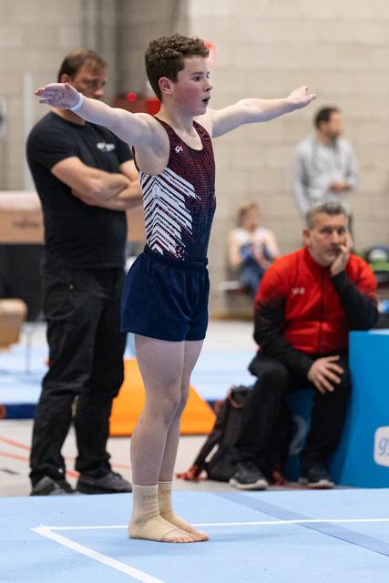 Young gymnast in burgundy and navy leotard stands in starting pose with arms extended on floor mat, coaches watching nearby
