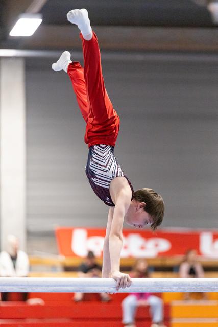 Young gymnast performs handstand on parallel bars wearing red top and patterned shorts in indoor training facility