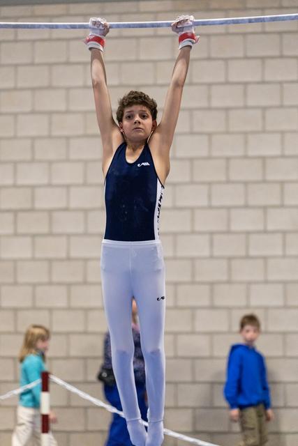 Young gymnast in navy leotard and white pants hangs from high bar with arms extended, focused expression in training gym