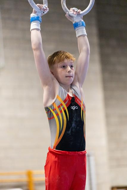 Young redhead gymnast hanging from rings with focused expression, wearing colorful leotard and red shorts in training hall