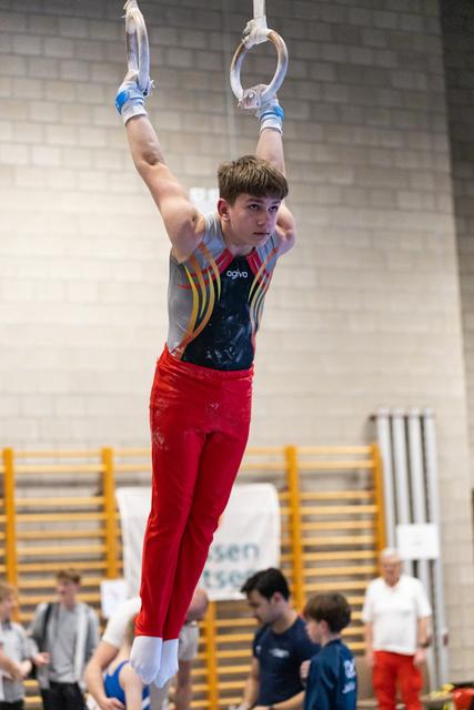 Young gymnast in red pants and colorful leotard hangs from still rings, focused expression, teammates visible below in gym