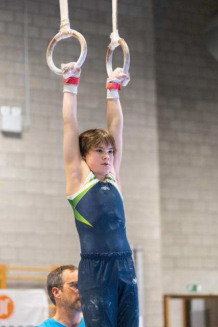 Young gymnast hanging from rings with focused expression while coach observes from below in training facility