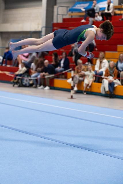 Young gymnast performing a horizontal flip above blue floor mat while spectators watch from colorful bleachers