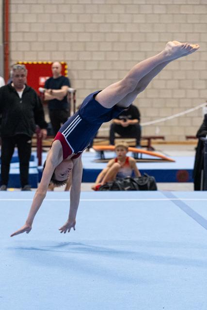 Young gymnast performs inverted handstand during floor routine while coaches and teammates watch in gym background