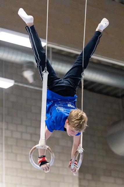Young gymnast performing inverted position on rings, arms extended upward, wearing blue shorts and black tracksuit jacket