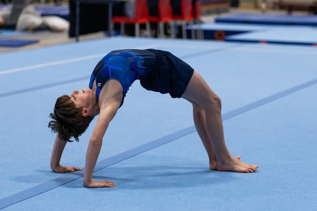 Young gymnast performs a backbend on the blue training mat, demonstrating flexibility and strength in her warm-up routine.