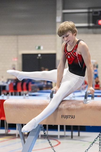 Young gymnast performing circles on Fujitsu pommel horse, displaying intense concentration in white pants and red-black leotard