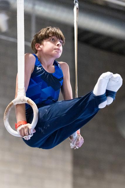 Young boy in blue leotard performs on gymnastics rings, looking up with focused expression while holding horizontal position