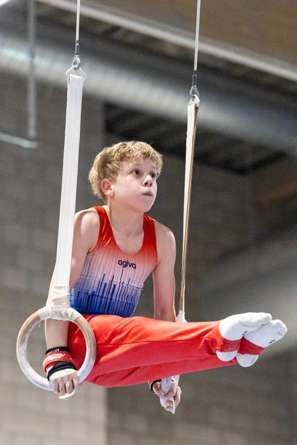 Young gymnast in red and blue gradient leotard demonstrates strength hold on rings, looking upward with focused expression
