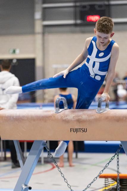 Young gymnast in blue unitard performs a leg extension on pommel horse, displaying concentration and form during routine