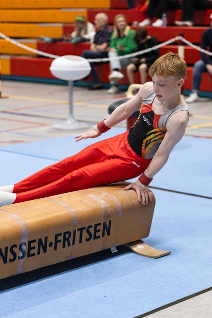 Young gymnast in red pants performs on pommel horse, displaying intense concentration during routine at indoor gymnastics event