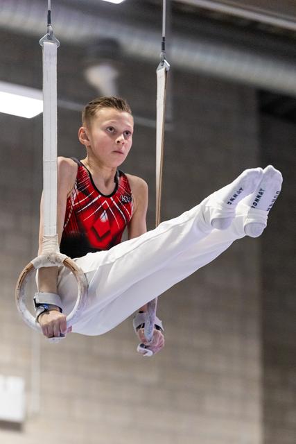 Young gymnast performs L-sit position on rings, displaying intense concentration and strength in red and black leotard