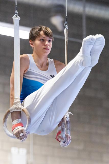 Young male gymnast performing rings routine with white pants and blue leotard, displaying focus and strength during training