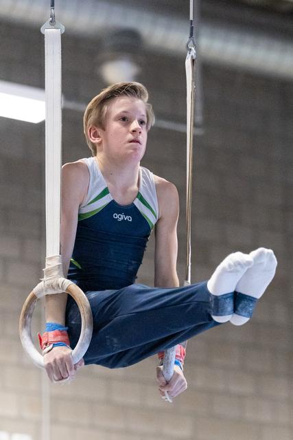 Young gymnast performing on rings with focused expression, wearing navy Ogiva uniform and white hand grips in training facility