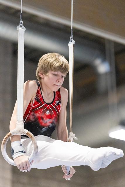 Young male gymnast performs on rings, concentrating intently on his form in white pants and red leotard