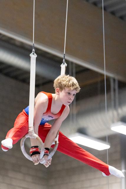 Young gymnast performing on rings with intense concentration, body extended horizontally in red and blue uniform