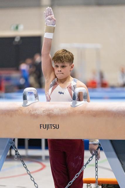 Young gymnast raises hand in salute while gripping pommel horse handles, displaying focus and determination during routine