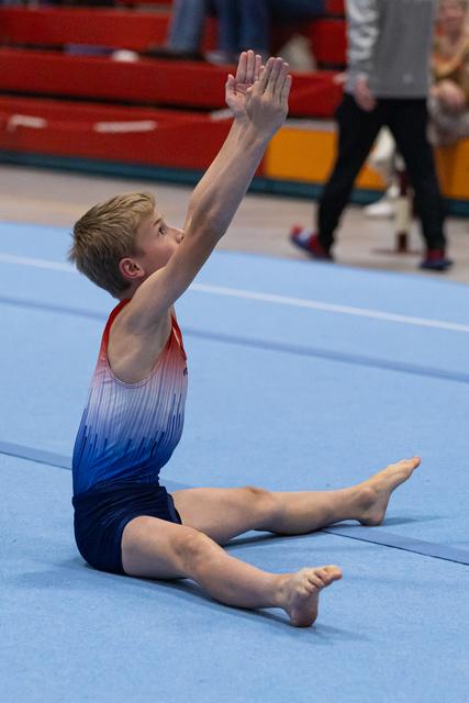 Young male gymnast performs seated position with arms raised on blue floor mat during training