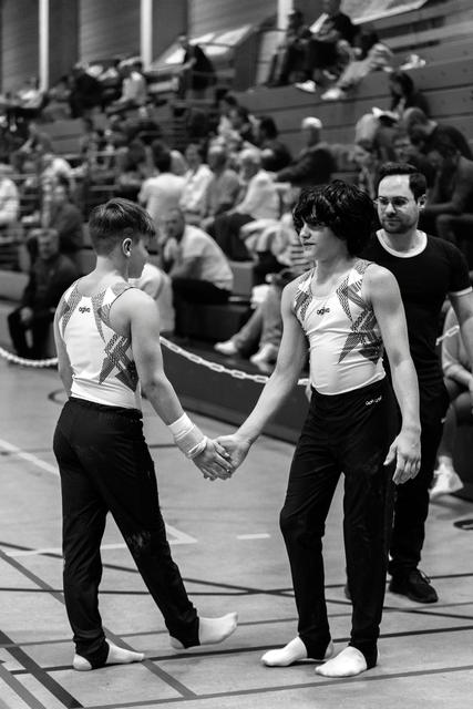 Two young gymnasts in matching uniforms shake hands on the competition floor, displaying good sportsmanship