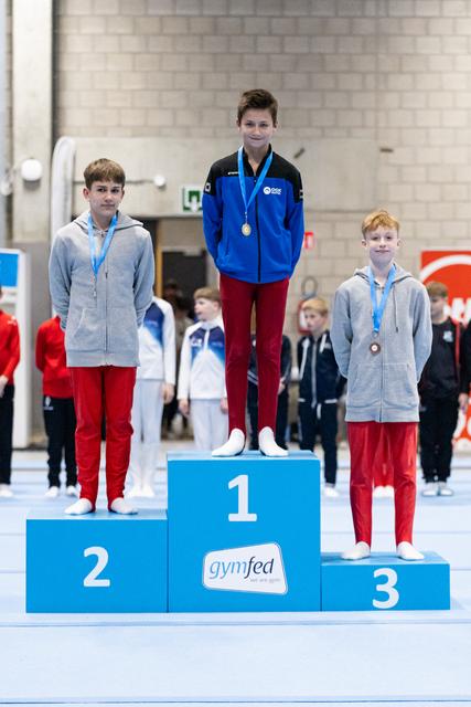 Three young gymnasts stand on award podium, winner in blue jacket on first place, two others in grey hoodies on second and third