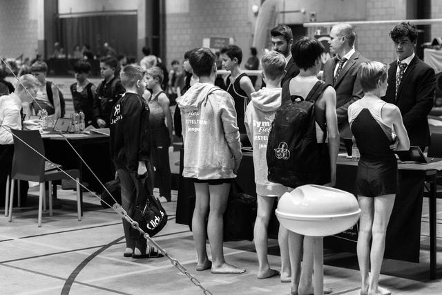 Young gymnasts stand in line wearing team vests, awaiting their turn by the equipment tables in a sports hall