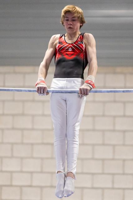 Young gymnast in red and black leotard performs on horizontal bar, focused expression, white pants, brick wall background