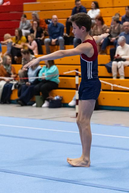 Young male gymnast in navy and maroon leotard stands barefoot in ready position on floor mat, arms extended, spectators watching