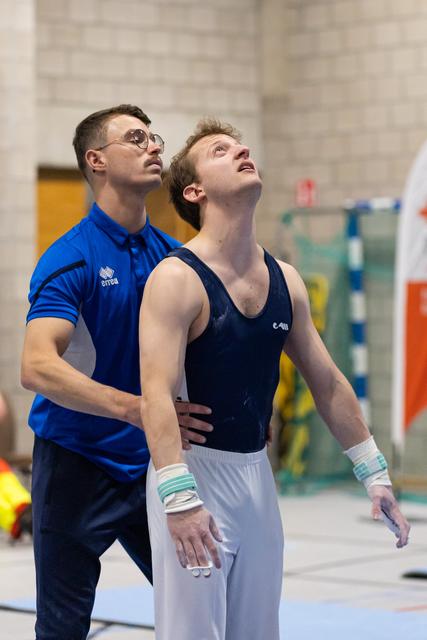 Coach in blue polo steadies a gymnast in navy leotard as they both gaze upward together in a training hall