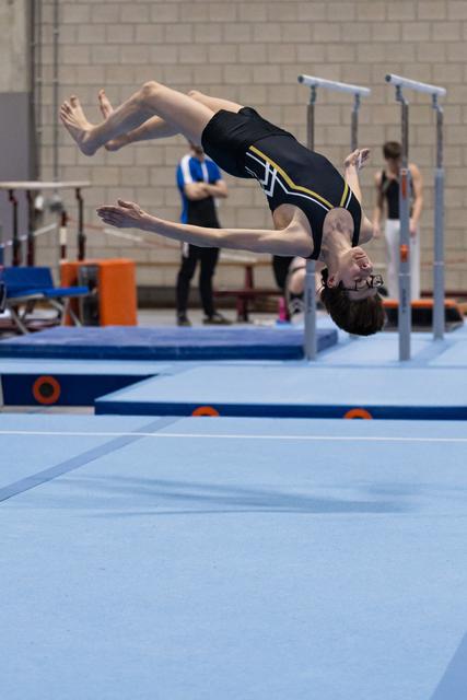 Gymnast performing a backflip mid-air above training mat, body arched in controlled rotation, training hall background
