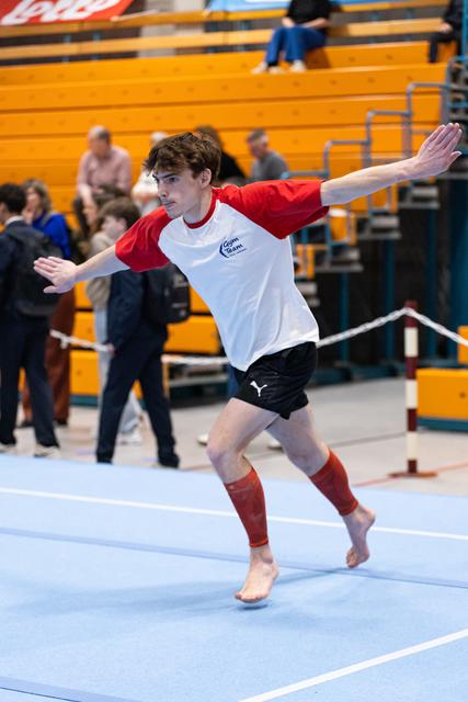 Young gymnast in red and white shirt performs a balance pose with extended arms on blue floor mat during indoor event