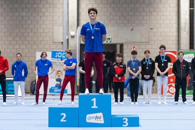 Young gymnast stands proudly on first place podium wearing gold medal, with blue jersey and competitors standing behind